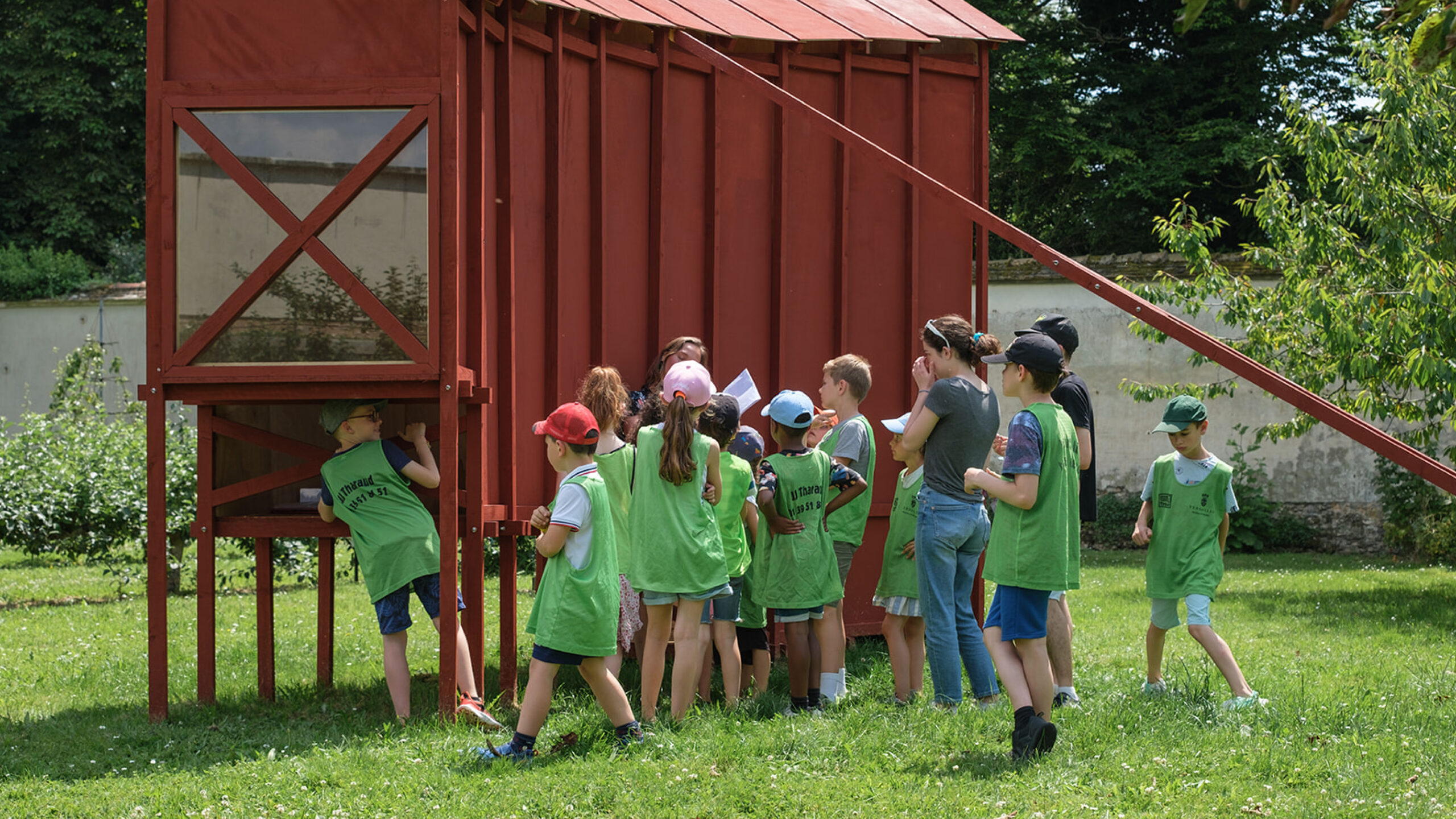 Groupe de jeunes enfants se trouvant dans les jardins de Versailles, observant une structure en bois rouge.