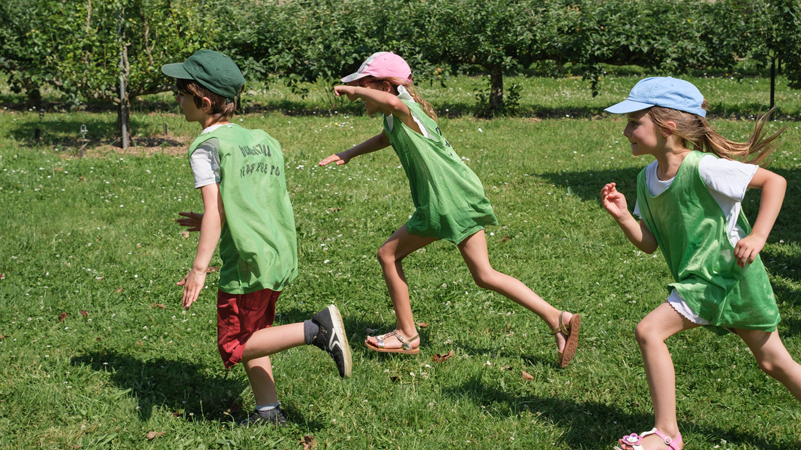 Trois jeunes enfants courant dans un jardin, entouré d'arbre fruitier.