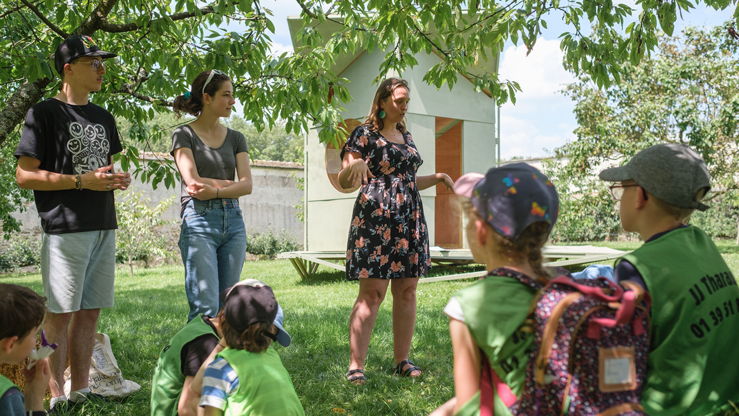 Un groupe d'enfants à l'ombre d'un arbre, écoutant une femme parler, en arrière-plan une structure de bois peint en blanc.