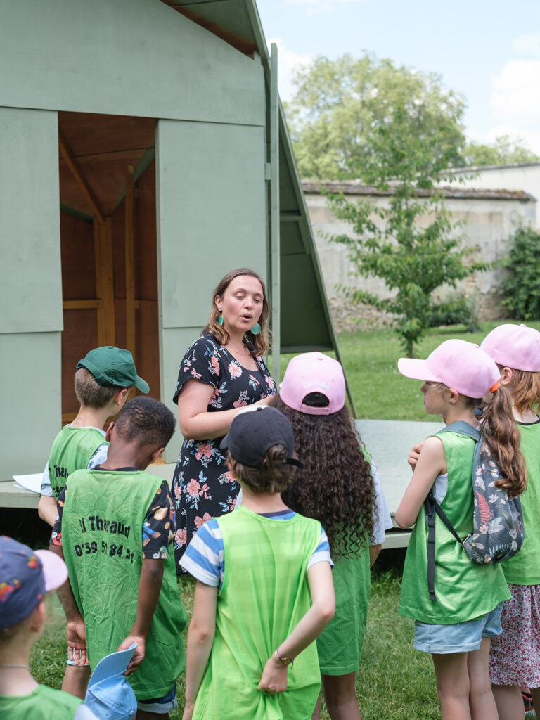 Un groupe d'enfants à l'ombre d'un arbre, écoutant une femme parler, en arrière-plan une structure de bois peint en vert.