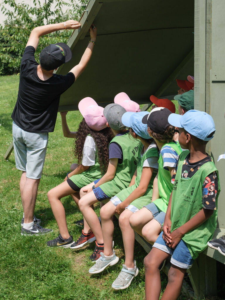 Un groupe de jeunes enfants assis sur une structure en bois peint en vert dans les jardins de Versailles, en train d'écouter un homme de dos parler.