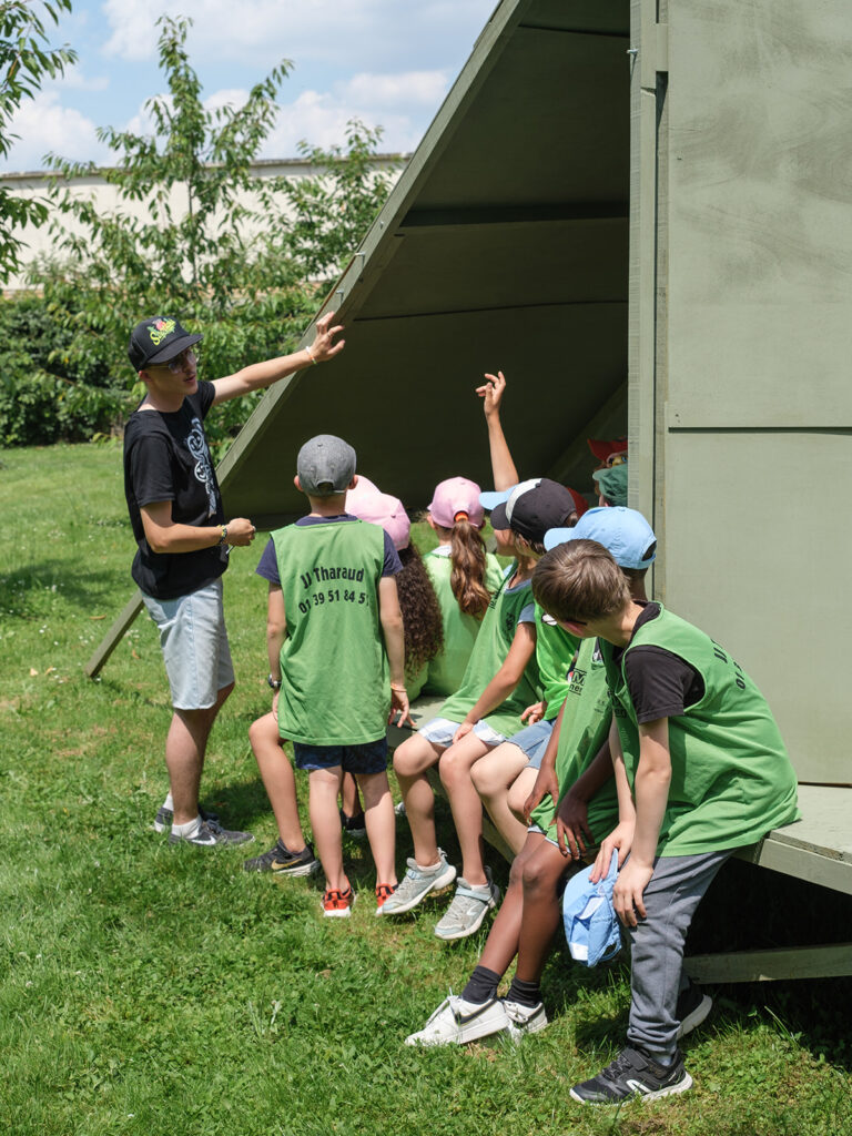 Un groupe de jeunes enfants assis sur une structure en bois peint en vert dans les jardins de Versailles, en train d'écouter un homme de parler.